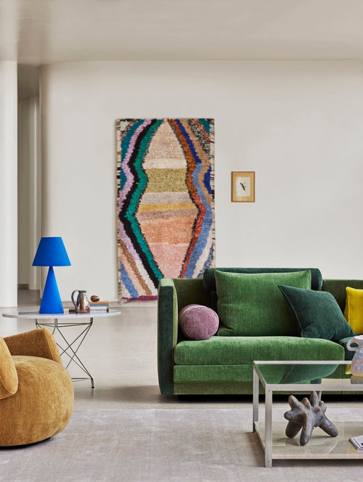 A modern living room featuring vibrant green velvet sofas with colorful cushions, a white coffee table with artistic sculpture, and a bold blue table lamp, showcasing contemporary Danish interior design.