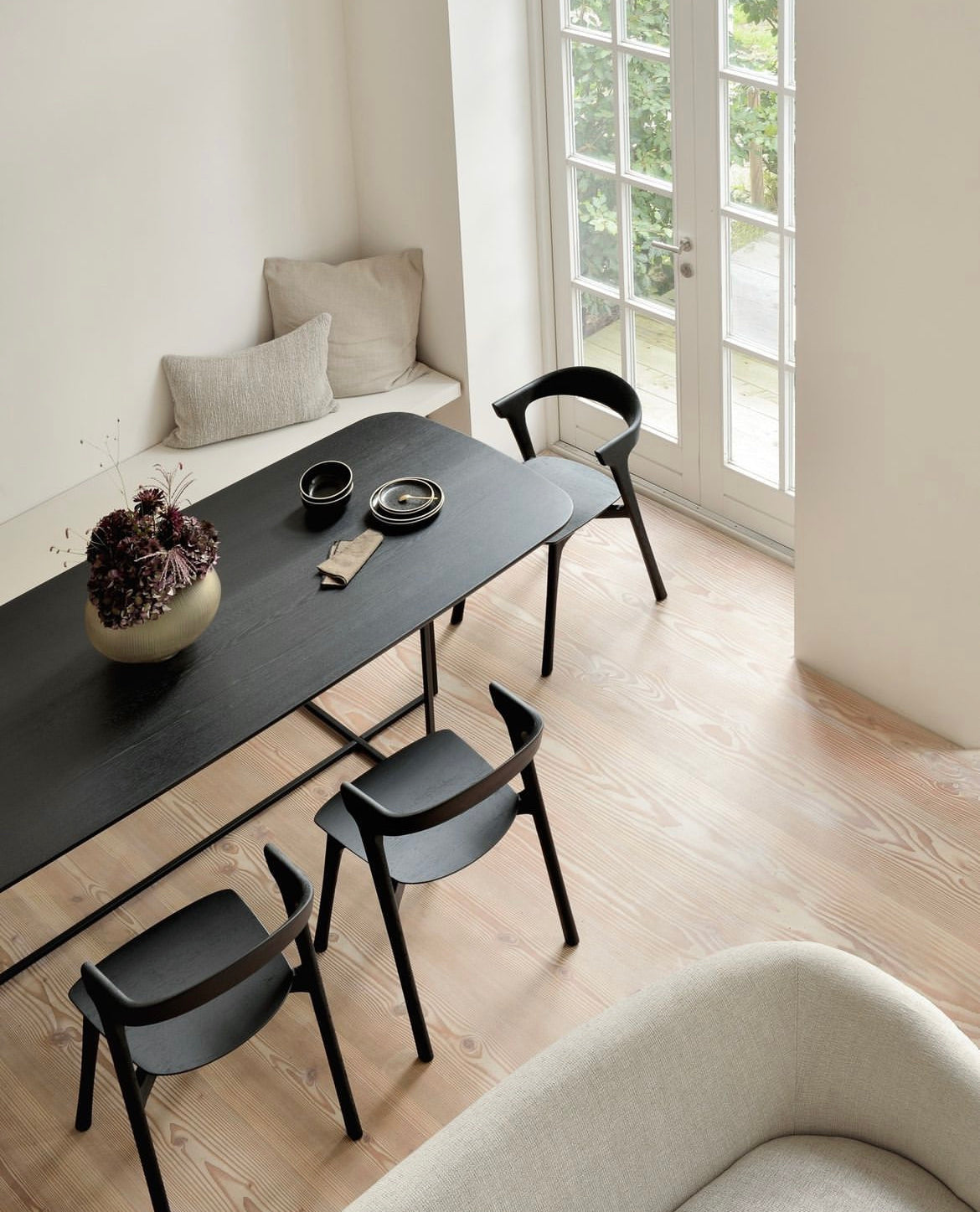 Minimalist dining area with light wood flooring, black dining table, black chairs, and a cozy window seat with neutral cushions, brightened by natural light.