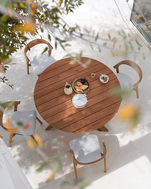 Aerial view of a round Ethnicraft wooden dining table with four modern chairs, set with breakfast items, in a bright, sunlit outdoor patio area.