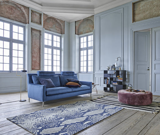 A spacious living room with large, historic windows, elegant white wall paneling, a plush blue sofa, a pink velvet ottoman, and a patterned blue and white rug, showcasing timeless design and durable furnishings.