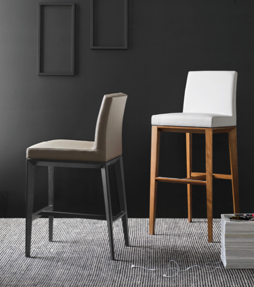 A modern interior with two different counter height stools: one with a beige upholstered seat and black wooden legs, and the other with a white upholstered seat and natural wood frame, set against a dark wall with minimal decor.