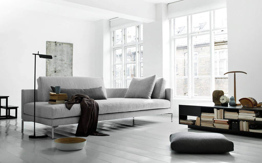 A minimalist Scandinavian living room featuring a light-colored Plano sofa, large windows with natural daylight, and modern decor elements.
