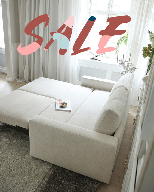 A modern, cream-colored sleeper sofa bed on sale in a bright, stylish living room with large windows and white curtains.