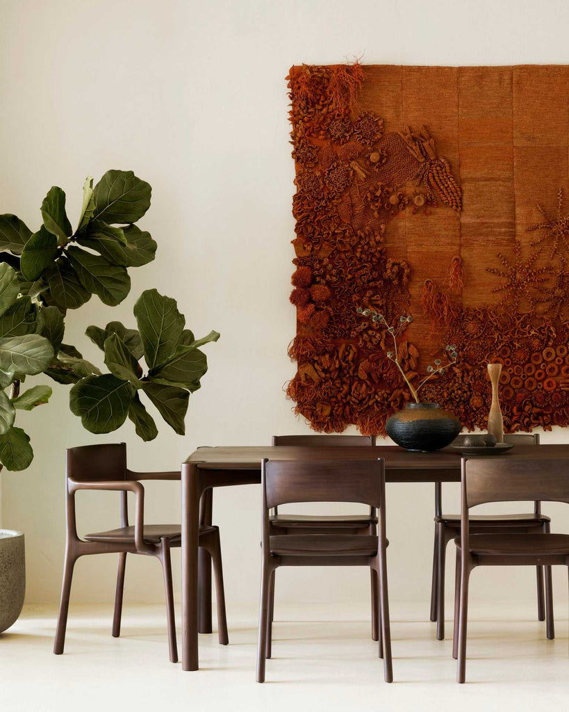 Elegant dining area featuring a dark wooden table, surrounded by matching chairs, with a textured rust-colored textile wall hanging and decorative vase