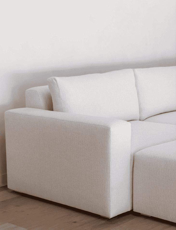 A close-up of a modern sectional sofa in a light beige textured fabric, showing the armrest, back cushion, and part of the seat against a plain white wall and light wood floor.