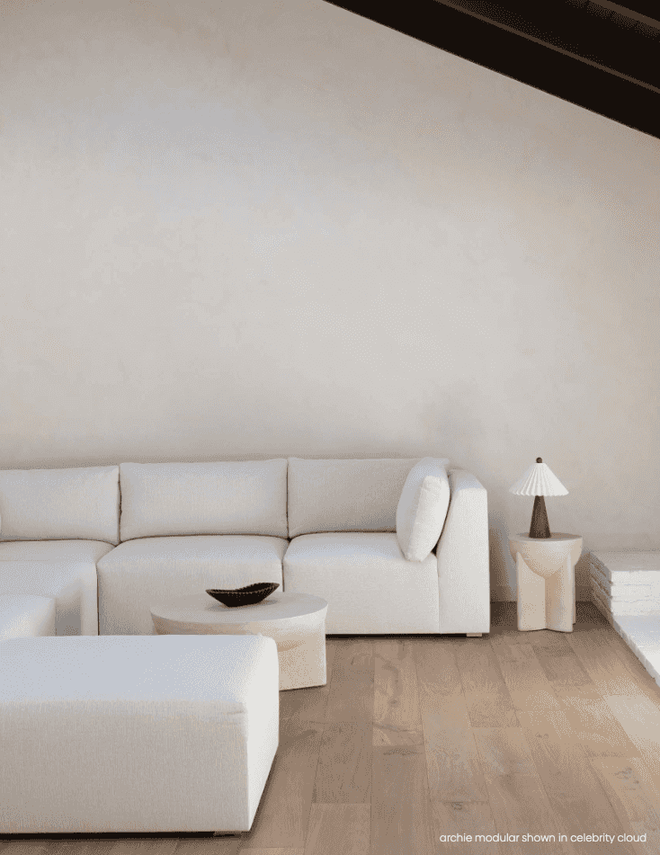 A modern light beige sectional sofa with matching ottomans in a minimalist, warm-toned living room setting with light wood flooring and a textured cream wall.