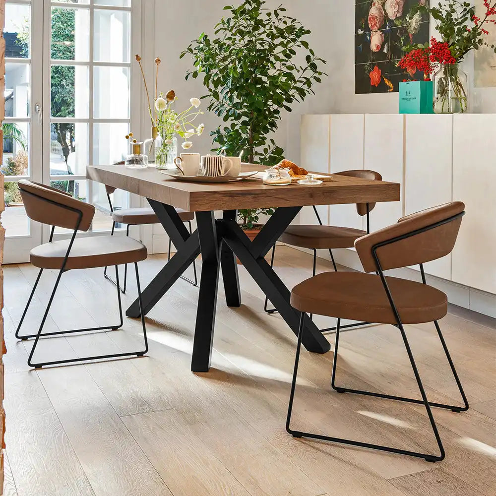Wooden dining table with a thick natural wood top and black metal X-shaped legs, set in a bright, cozy kitchen with plants