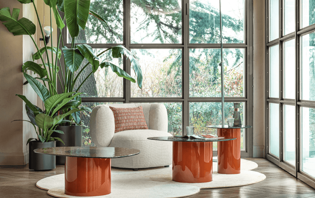 Living room with large windows, green plants, and a cream upholstered sofa with a rust-colored pillow, featuring three glossy orange cylindrical coffee tables with black marble tops, on a white rug.