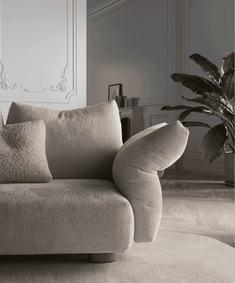 Plus Sofa by Art Nova in a neutral beige fabric with plush cushions, set in a stylish living room with decorative wall paneling and a large green plant
