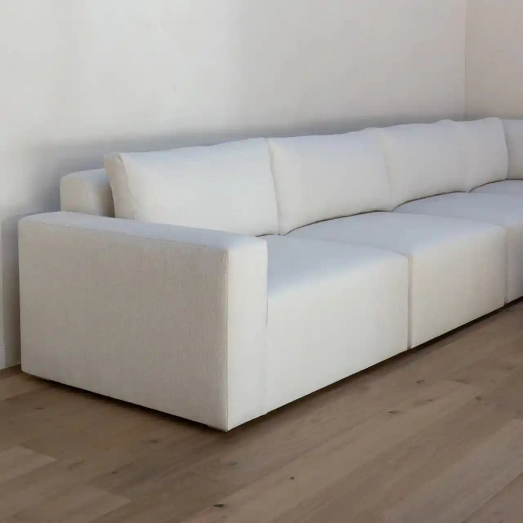 A modern modular white fabric sofa with a low, boxy silhouette, set against a minimalist white wall and light wood flooring.