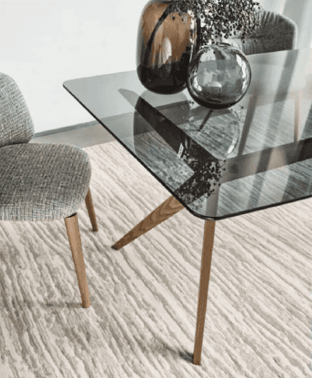 Dining table with a glass top and wooden legs, decorated with vases and placed on a textured rug in a stylish living space.