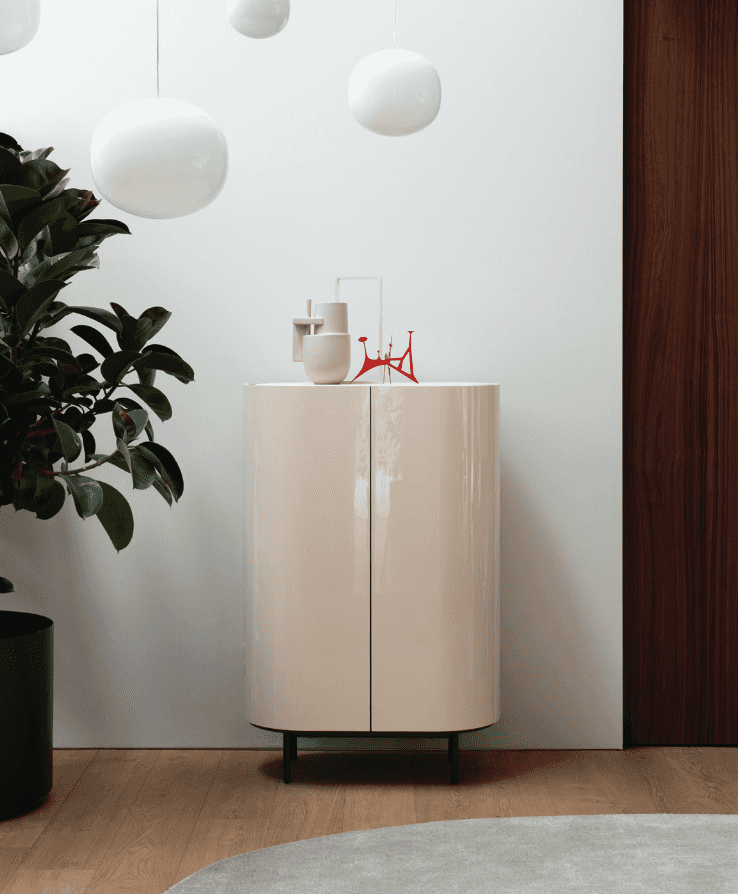 Elegant white storage cabinet with a glossy surface, rounded doors, and black legs, set against a light wall with hanging white pendant lights and decorative items on top