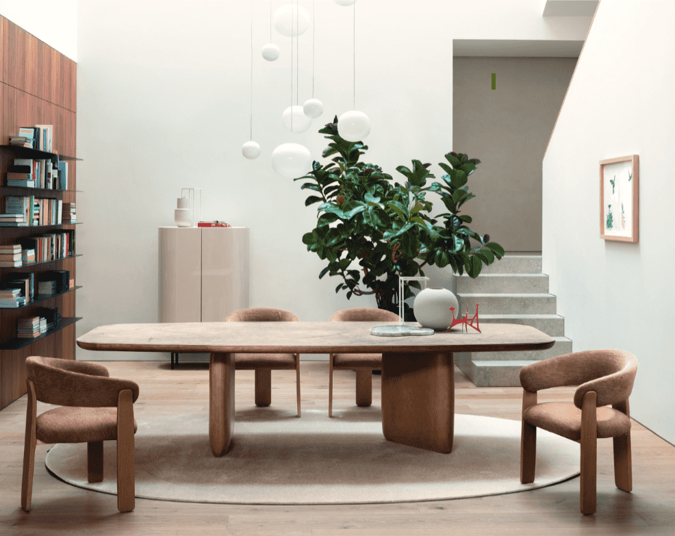 Elegant Riverstone elliptical edge dining table with wooden base in a modern dining room, surrounded by beige upholstered chairs, with a large green plant and minimalist decor