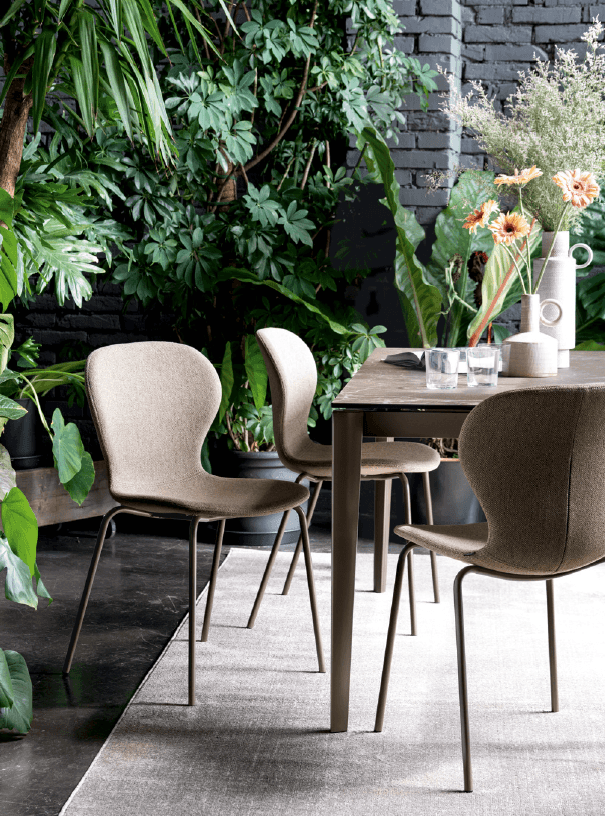 Set of beige upholstered dining chairs with curved backrests and slender metal legs, arranged around a wooden table in a lush indoor garden setting