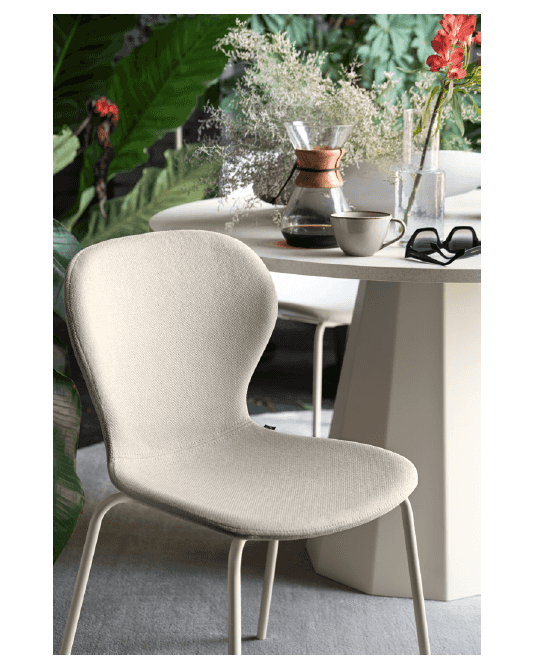 Cream-colored fabric dining chair with rounded backrest and metal legs, positioned near a white table with decorative vases and plants in a bright room