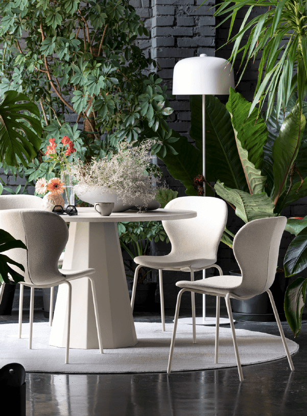 Indoor dining area with a round white table surrounded by four beige upholstered chairs, lush green plants, and a white floor lamp