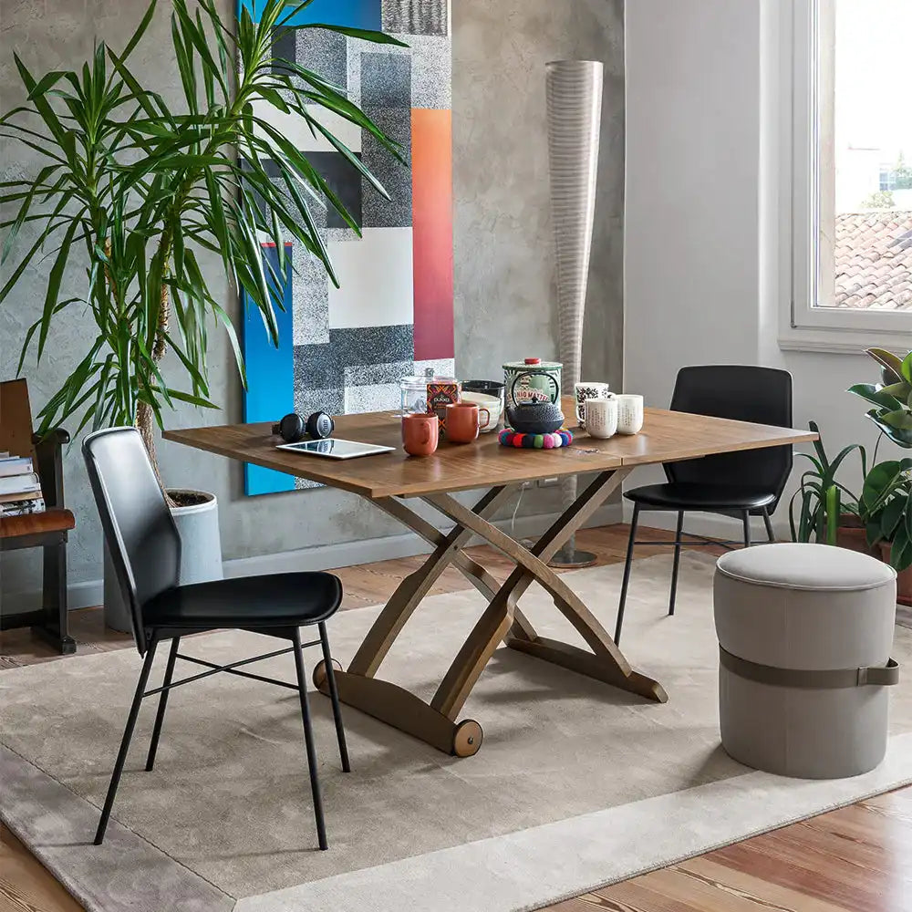 Wooden multi-purpose dining table with X-shaped base in a modern living room, surrounded by black and beige chairs, with decorative items on top.