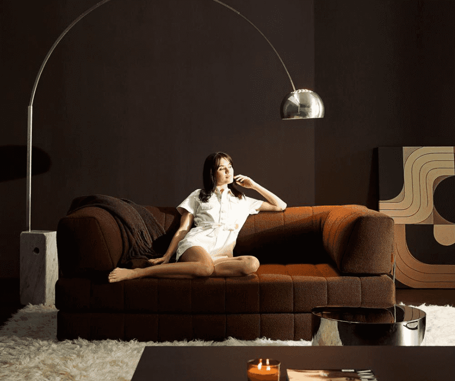 Woman sitting on a brown sofa in a dimly lit room with a large floor lamp.