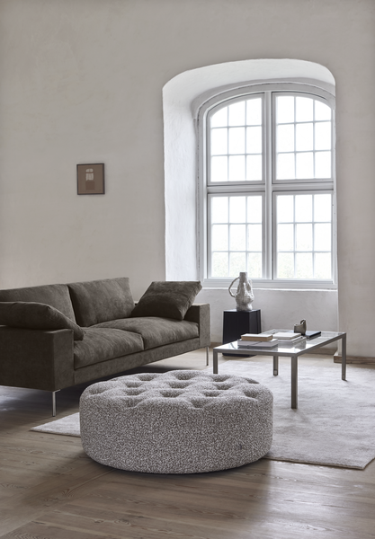 Shu Sofa by Eilersen in a cozy corner with a round tufted ottoman and modern decor, ideal for stylish and comfortable living rooms.