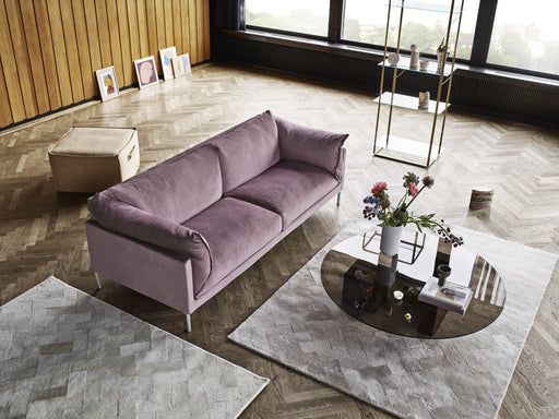 A stylish living room featuring a plush purple sofa, modern glass and gold shelving unit, abstract artwork, and a floral arrangement on a black coffee table, with large windows and wooden wall paneling.