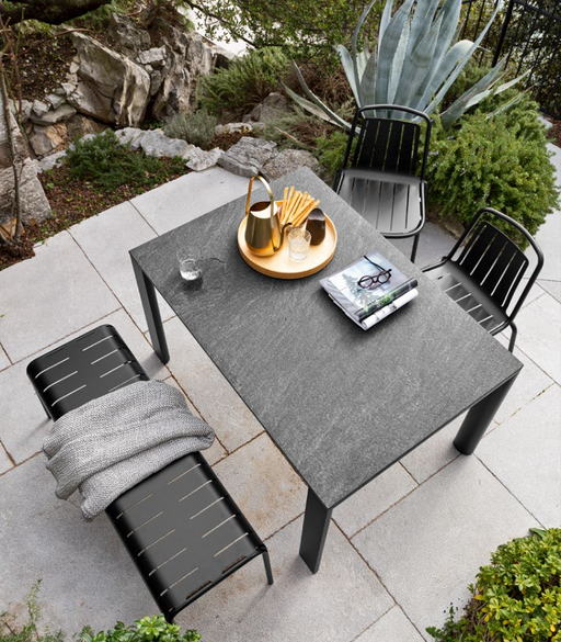 A modern outdoor dining setup featuring Connubia Italian furniture with a sleek black table, matching chairs, and a bench, surrounded by lush greenery and rocks.
