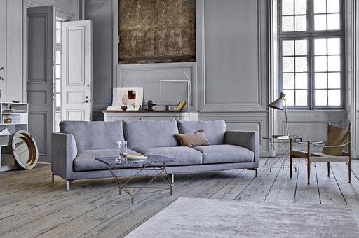 Elegant Scandinavian living room featuring a light gray sofa, minimalist glass coffee table, and vintage-inspired wooden chairs with a modern brass desk lamp, showcasing Danish-Japanese furniture design collaboration.