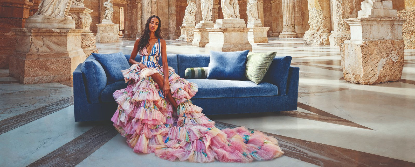 A fashion model wearing a colorful, ruffled gown sitting on a deep blue velvet sofa in an ancient Greek-style marble hall with classical statues and columns, highlighting vibrant color trends for 2027.