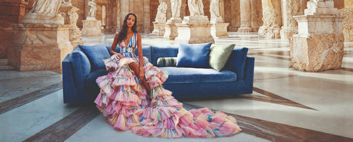 A fashion model wearing a colorful, ruffled gown sitting on a deep blue velvet sofa in an ancient Greek-style marble hall with classical statues and columns, highlighting vibrant color trends for 2027.