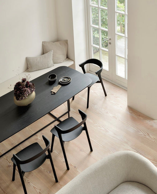 Minimalist dining area with light wood flooring, black dining table, black chairs, and a cozy window seat with neutral cushions, brightened by natural light.