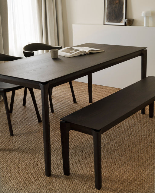 A modern, minimalist dining room setup featuring a dark wooden dining table, matching bench, and black chairs, with natural light filtering through sheer curtains.