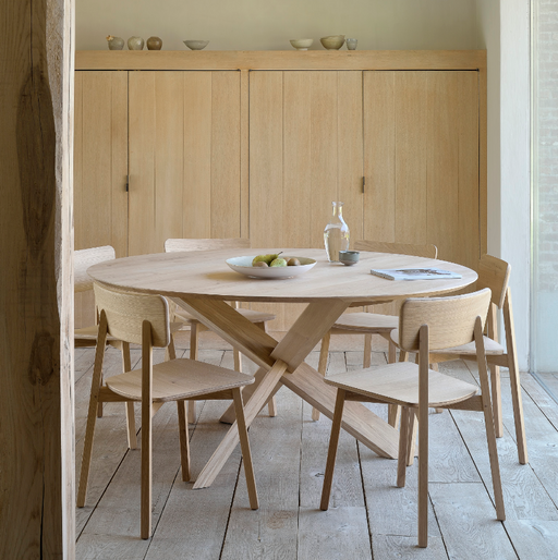 A minimalist dining room featuring light wood furniture, including a round table with a unique crossed-leg design, surrounded by matching wooden chairs, with a wooden cabinet in the background, emphasizing natural wood finishes and indoor air quality.