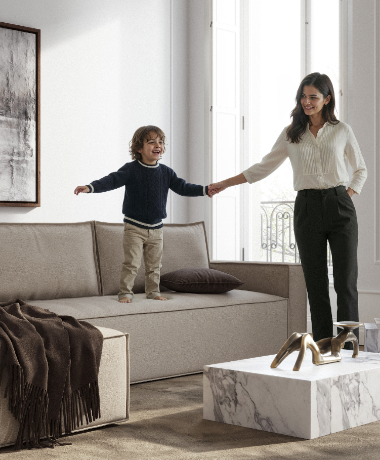 A happy woman and young boy playing on a beige sofa in a bright, modern living room with large windows, stylish decor, and a marble coffee table.