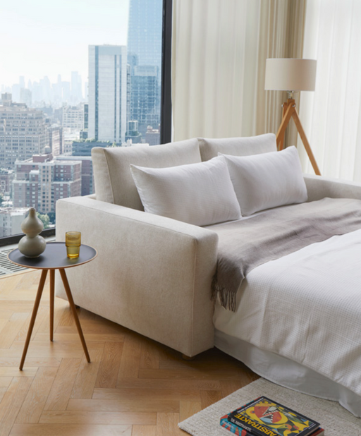 A modern living room featuring a sleek sofa bed with plush white pillows, large window with city skyline view, and a stylish side table with decorative vases and a glass, highlighting the best-reviewed Innovation Living sofa bed.