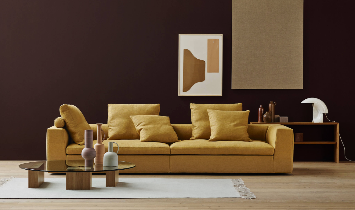 A modern home office or living room setup with a mustard yellow sofa, decorative pillows, abstract wall art, and a sleek glass coffee table, illustrating the balance of life and work from home in the new normal.