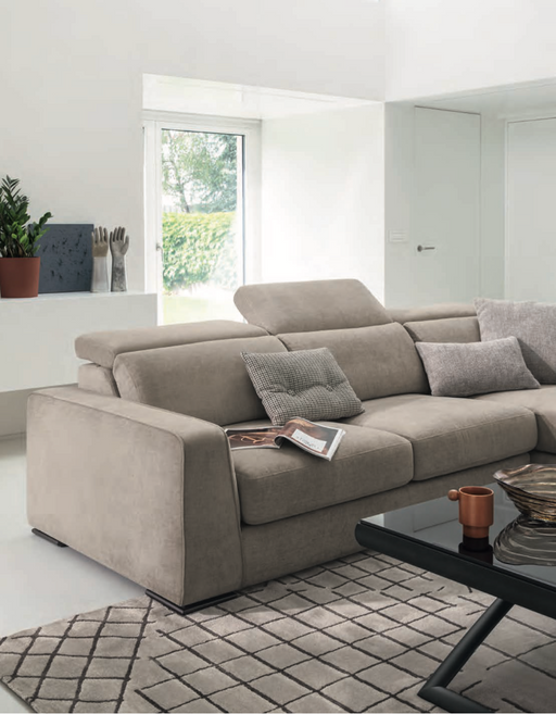 A modern, beige fabric sofa with adjustable headrests in a bright, minimalist living room featuring a black coffee table, patterned rug, and large window with greenery outside.