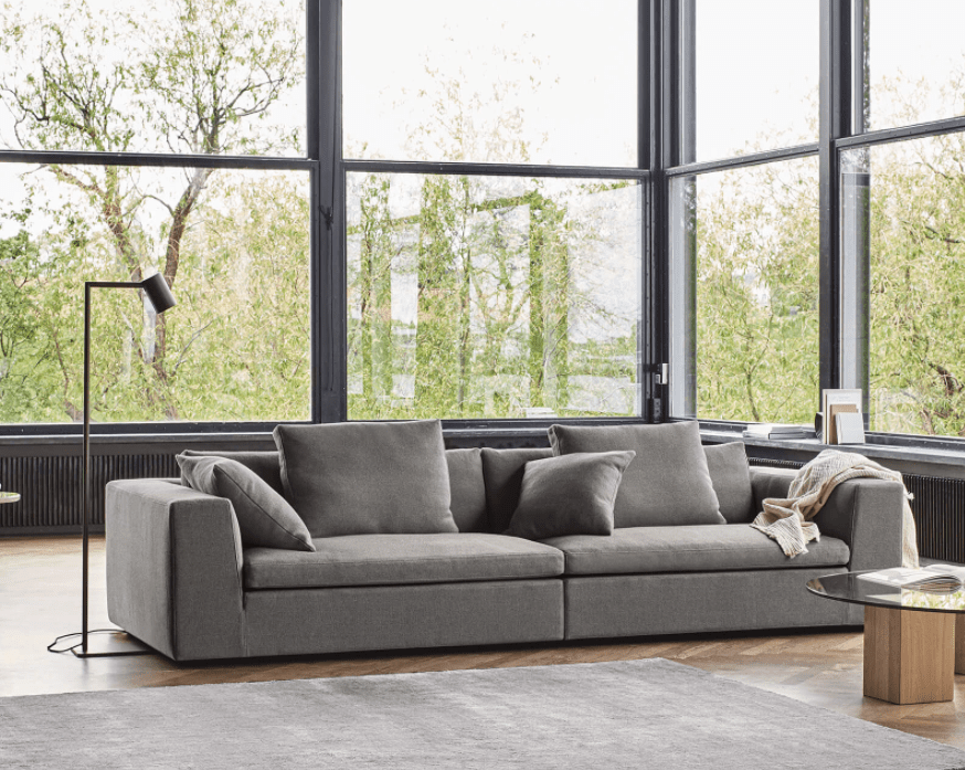 Modern gray Eilersen quick ship sofa with plush cushions in a bright living room featuring large floor-to-ceiling windows and minimalist decor.