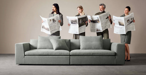 A group of four diverse adults reading newspapers while standing behind a modern, minimalist light gray sofa in a neutral-toned room, showcasing timeless Danish design.