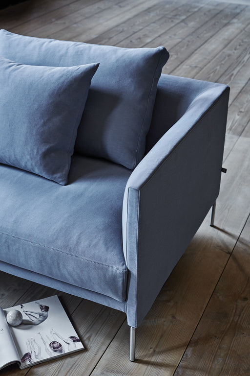 A modern, light blue velvet sofa with sleek metal legs, featuring plush cushions and a contemporary design, set on a wooden floor with an open magazine nearby.