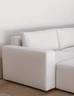 A close-up of a modern sectional sofa in a light beige textured fabric, showing the armrest, back cushion, and part of the seat against a plain white wall and light wood floor.