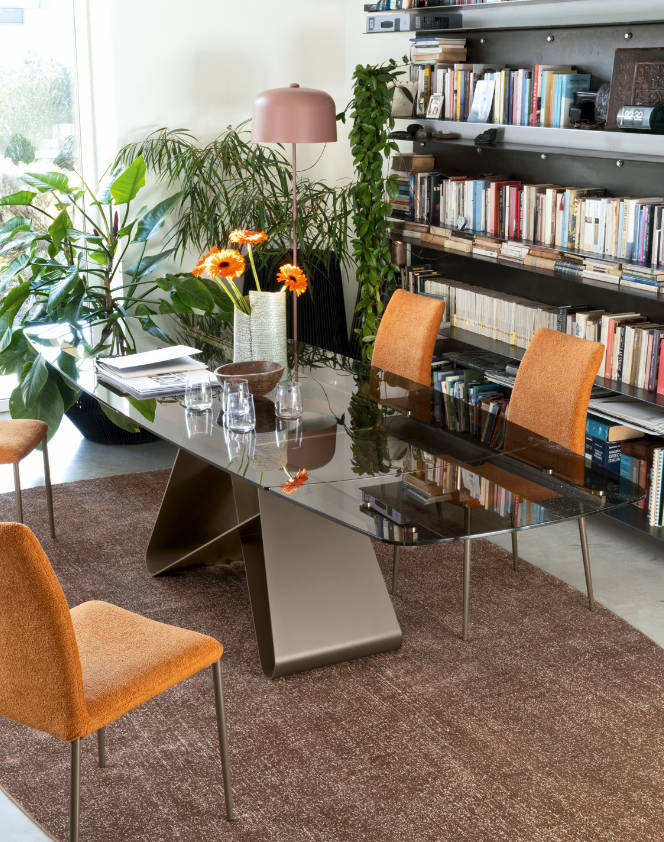 A modern dining room scene featuring a glass-top table with a sculptural metallic base, surrounded by three orange upholstered chairs on a brown rug. The room is styled with potted plants, a pink floor lamp, and a large bookshelf filled with books.
