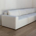 A modern modular white fabric sofa with a low, boxy silhouette, set against a minimalist white wall and light wood flooring.