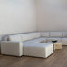 A modern light beige sectional sofa with matching ottomans in a minimalist, warm-toned living room setting with light wood flooring and a textured cream wall.